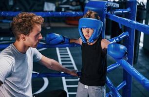 Der junge Boxtrainer hilft dem kleinen Jungen in Schutzkleidung zwischen den Runden am Ring foto