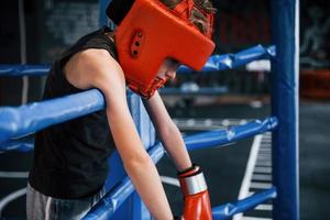 Müder Junge in Schutzausrüstung, der sich auf die Knoten des Boxrings stützt foto