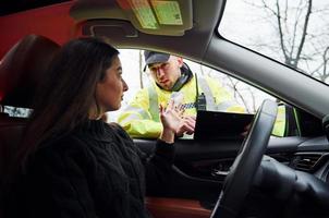 männlicher polizist in grüner uniform, der fahrzeug auf der straße überprüft. Frau, die versucht, Bestechungsgelder zu geben foto