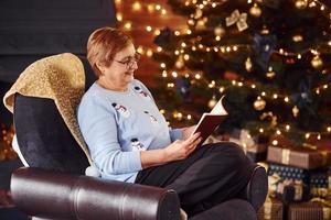 alte frau, die auf dem stuhl mit buch im festlichen neujahrsraum mit weihnachtsdekorationen sitzt foto