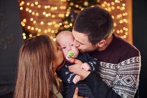 vater und mutter küssen ihr kind im weihnachtlich dekorierten zimmer foto