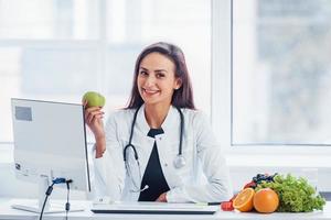 Ernährungsberaterin im weißen Kittel sitzt drinnen im Büro am Arbeitsplatz mit Apfel in der Hand foto
