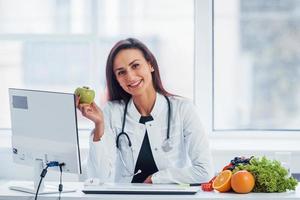 Ernährungsberaterin im weißen Kittel sitzt drinnen im Büro am Arbeitsplatz mit Apfel in der Hand foto