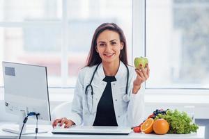 Ernährungsberaterin im weißen Kittel sitzt drinnen im Büro am Arbeitsplatz mit Apfel in der Hand foto