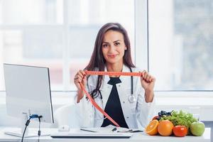 Ernährungsberaterin im weißen Kittel sitzt drinnen im Büro am Arbeitsplatz mit Maßband in den Händen foto