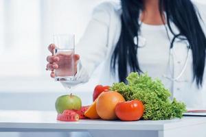 weibliche Ernährungsberaterin im weißen Kittel, die drinnen im Büro am Arbeitsplatz mit einem Glas Getränk sitzt foto