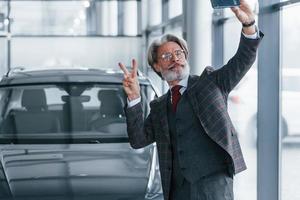 positiver älterer mann mit grauen haaren, der selfie gegen modernes auto drinnen macht foto