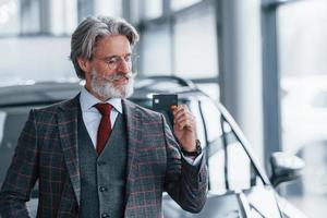 Mann mit grauem Haar und Schnurrbart, der drinnen mit Kreditkarte in der Hand gegen ein modernes Auto steht foto