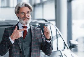 Mann mit grauem Haar und Schnurrbart, der drinnen mit Kreditkarte in der Hand gegen ein modernes Auto steht foto