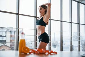 Frau mit sportlichem Körper, die drinnen neben einem Tisch mit Orangensaft und Maßband steht foto