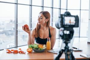 Vloggerin mit sportlichem Körper, die drinnen neben einem Tisch mit gesundem Essen steht