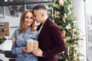 Schönes Paar, das zur Neujahrszeit in der Nähe von Auto und Weihnachtsbaum steht und Überraschung macht, indem es eine Geschenkbox gibt foto