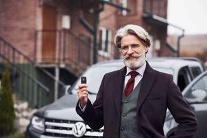 Modischer älterer Mann mit grauem Haar und Bart, der draußen auf der Straße in der Nähe seines Autos mit Schlüsseln in der Hand steht foto