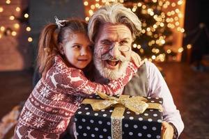 Fröhlicher, altmodischer Senior mit grauem Haar und Bart, der mit einem kleinen Mädchen in einem geschmückten Weihnachtszimmer sitzt foto