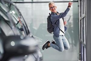 Fröhlicher Hipster-Typ, der drinnen vor grauem Hintergrund und in der Nähe des Autos springt foto