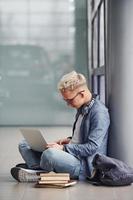 junger Hipster, der drinnen in der Nähe von grauem Hintergrund mit Laptop und Büchern sitzt foto