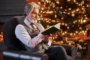 Porträt eines stilvollen Seniors mit grauem Haar und Bart, der ein Buch in einem dekorierten Weihnachtszimmer liest foto