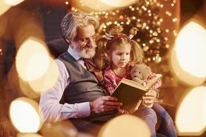 Fröhlicher, altmodischer Senior mit grauem Haar und Bart, der mit einem kleinen Mädchen in einem geschmückten Weihnachtszimmer sitzt und ein Buch liest foto