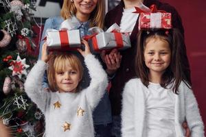 Zwei kleine Mädchen stehen zusammen mit Mama und Papa im Zimmer mit Weihnachtsbaum mit Geschenkboxen foto