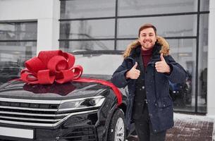 glücklicher besitzer eines neuen modernen autos, das draußen steht und lächelt. rote geschenklänge auf dem auto foto