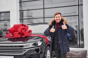 glücklicher besitzer eines neuen modernen autos, das draußen steht und lächelt. rote geschenklänge auf dem auto foto