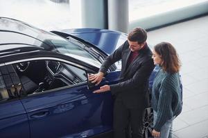 professioneller verkäufer, der junges mädchen unterstützt, indem er drinnen ein neues modernes auto auswählt foto