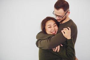 glückliches multiethnisches Paar in Freizeitkleidung, das sich drinnen im Studio umarmt. Kaukasier mit asiatischer Freundin foto