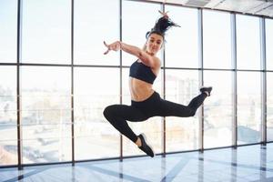 Junge sportliche Frau in Sportbekleidung springt und macht sportliche Tricks gegen Fenster in der Luft foto