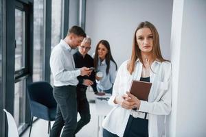 porträt einer fröhlichen blondine mit notizblock, die vor einem jungen erfolgreichen team steht, das drinnen im büro zusammenarbeitet und kommuniziert foto