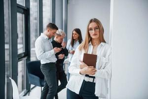 porträt einer fröhlichen blondine mit notizblock, die vor einem jungen erfolgreichen team steht, das drinnen im büro zusammenarbeitet und kommuniziert foto