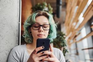 alternatives mädchen mit brille und grünen haaren, die tagsüber mit telefon in den händen drinnen sitzen foto