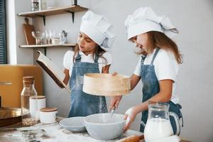 Zwei kleine Mädchen in blauer Kochuniform bereiten Essen zu, indem sie ein Sieb in der Küche verwenden und das Quittungsbuch lesen foto
