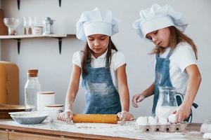 Zwei kleine Mädchen in blauer Kochuniform kneten Teig in der Küche foto