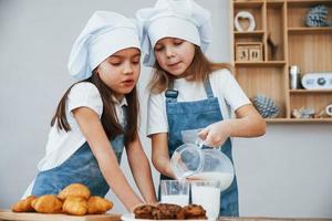 Zwei kleine Mädchen in blauer Kochuniform gießen Milch in Gläser in der Küche mit Keksen auf dem Tisch foto