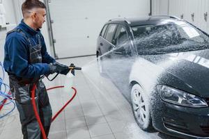 männlicher arbeiter in uniform, der neues modernes auto wäscht, das mit seife bedeckt ist. Konzeption des Dienstes foto