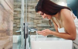 Schöne junge Frau, die im Badezimmer neben dem Spiegel steht und ihr Gesicht wäscht foto