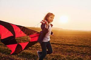 glückliches kleines Mädchen, das zur Sonnenaufgangszeit mit Drachen in den Händen auf dem schönen Feld läuft
