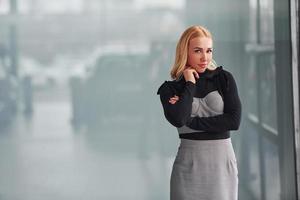 Porträt einer schönen Blondine in eleganter Kleidung, die drinnen vor grauem Hintergrund steht foto