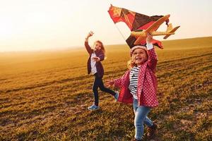 Zwei kleine Freundinnen amüsieren sich bei sonnigem Tag zusammen mit Drachen und Spielzeugflugzeug auf dem Feld foto