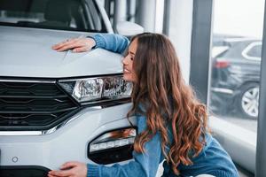 positive Frau im blauen Hemd, die ihr brandneues Auto umarmt. im Autosalon foto