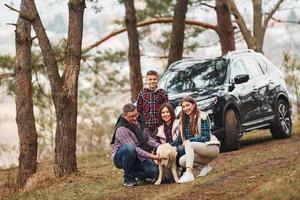 glückliche familie, die mit ihrem hund in der nähe eines modernen autos draußen im wald sitzt und spaß hat foto