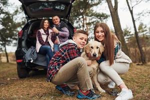 glückliche familie, die mit ihrem hund in der nähe eines modernen autos draußen im wald sitzt und spaß hat foto