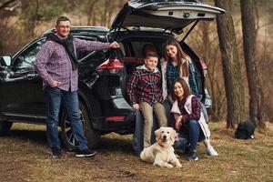 glückliche familie hat spaß mit ihrem hund in der nähe eines modernen autos im freien im wald foto