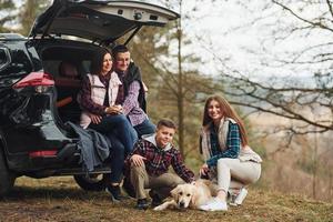 glückliche familie, die mit ihrem hund in der nähe eines modernen autos draußen im wald sitzt und spaß hat foto
