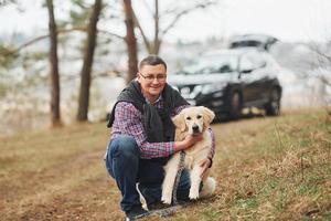 mann mit brille geht mit seinem hund draußen im wald spazieren. modernes schwarzes Auto dahinter foto