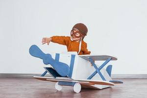 Kleiner Junge in Retro-Pilotenuniform, der sich im Innenbereich mit Spielzeugflugzeug amüsiert foto