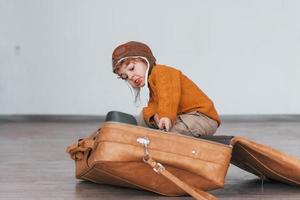 kleine jungen im retro-pilotenkostüm haben spaß und sitzen tagsüber im koffer drinnen foto