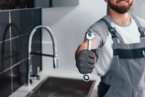 Nahaufnahme eines jungen professionellen Klempners in grauer Uniform, der einen Schraubenschlüssel in der Hand in der Küche hält foto