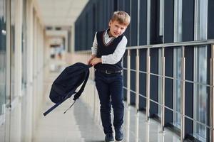 kleiner Schuljunge in Uniform, der mit Rucksack im Korridor spazieren geht foto