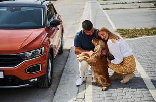 setzt sich mit haustier. Schönes Paar macht einen Spaziergang zusammen mit Hund im Freien in der Nähe des Autos foto